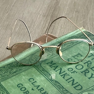 May include: Vintage gold-rimmed round glasses with clear lenses resting on a green book titled "The Story of Mankind" by Hendrik Willem van Loon.