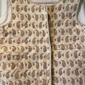 May include: A sleeveless quilted vest in an off-white color featuring a repeating paisley pattern in shades of red, blue, and green. The garment has a round neckline and is finished with a light brown and white striped trim along the edges.
