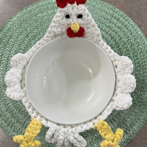 May include: White crocheted chicken bowl cosy with yellow feet and a red comb. The cosy is holding a white bowl.