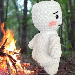Peut inclure: Peluche en crochet blanc avec une expression boudeuse et des joues roses. Le jouet est debout devant un feu de camp dans un décor de forêt.