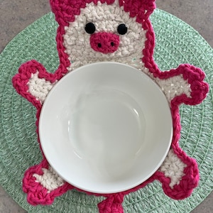 Puede incluir: Un calientaplatos de crochet rosa y blanco con forma de cerdo. El calientaplatos tiene forma de cerdo con ojos negros y un hocico rosa. El calientaplatos está sobre un mantel individual verde.
