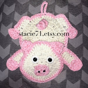 May include: A crocheted pink and white pig pot holder with black eyes and a pink nose. The pot holder has a loop for hanging. The text "stacie71.etsy.com" is visible on the pot holder.