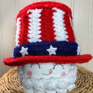 May include: A patriotic Uncle Sam-themed ceramic mug with a crocheted hat. The hat features red and white stripes, a blue band with star-shaped buttons, and a red brim. The mug has a smiling face with a white crocheted beard.