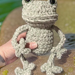 May include: A crocheted green frog plush toy with large, sparkly eyes. The frog is sitting on a rock with its legs outstretched.