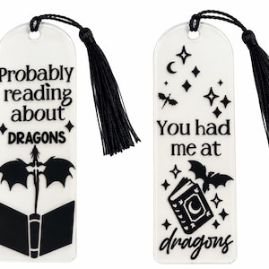 May include: Two translucent white bookmarks with black text and graphics. The left bookmark reads "Probably reading about DRAGONS" with a dragon and book icon. The right reads "You had me at dragons" with a book, dragon, and stars. Both have black tassels.