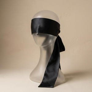 Puede incluir: Un antifaz de satén negro liso con una banda de tela ancha y suave que envuelve la cabeza y se asegura con lazos largos y fluidos que caen por el costado. El material tiene un brillo sutil que refleja la luz sobre su superficie.