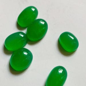 May include: Six smooth, oval-shaped green gemstones with a polished, cabochon finish. Each stone displays a vibrant, translucent emerald green hue with subtle internal variations and a reflective surface, arranged in a loose cluster.
