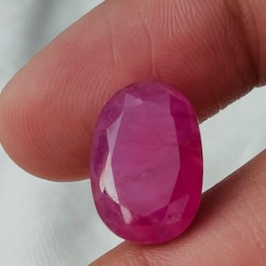 May include: A vibrant, oval-shaped, faceted gemstone with a deep magenta-pink hue. The stone features a polished surface with multiple geometric facets that reflect light, showcasing its translucent quality and saturated colour profile.