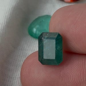 May include: A rectangular emerald-cut gemstone with a deep green colour and faceted surface. The stone is held in place, displaying its clear, polished edges and internal structure. In the background, a smaller green stone and a red stone are visible.