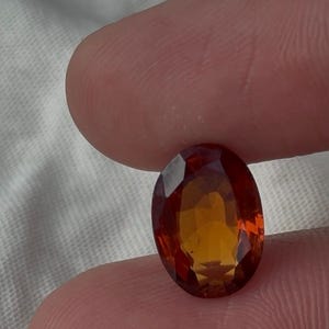 May include: An oval-cut gemstone featuring a vibrant gradient of colours, transitioning from a deep, saturated reddish-orange at the edges to a bright, warm golden-yellow in the centre, with a faceted surface that reflects light across its polished facets.