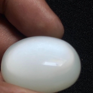 May include: An oval-shaped, smooth, and polished gemstone with a translucent, milky white appearance. The stone features a rounded cabochon cut that reflects light across its surface, displaying a soft, glowing sheen throughout the entire piece.