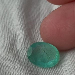 May include: An oval-shaped, faceted emerald gemstone with a vibrant green hue and visible internal inclusions. The gemstone features a polished surface with multiple geometric facets that reflect light, showcasing its translucent quality and deep colour.
