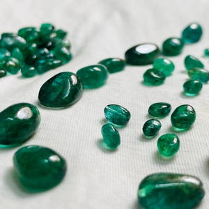 May include: A collection of loose, polished emerald gemstones in various sizes and shapes. The stones exhibit a deep, vibrant green colour with translucent qualities and smooth, rounded surfaces, scattered across a white textured fabric surface.