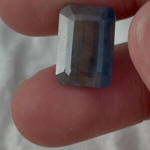 May include: A rectangular, emerald-cut gemstone with a deep blue and greyish-purple hue. The stone features faceted surfaces that reflect light, displaying a translucent quality with visible internal colour variations across its polished surface.