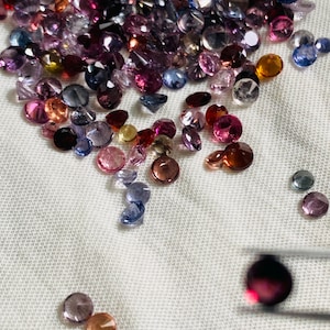 May include: A collection of loose, faceted gemstones in various colours including shades of pink, purple, blue, orange, and red. The stones are round and reflective. A pair of metal tweezers holds a single dark red gemstone in the lower right corner.