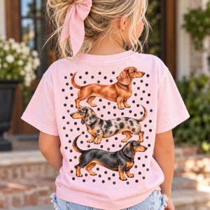 Puede incluir: Una camiseta rosa claro con un estampado gráfico de tres perros salchicha de diferentes colores de pelaje—marrón sólido, moteado y negro con fuego—dispuestos verticalmente sobre un patrón de fondo de pequeños puntos negros espaciados uniformemente.