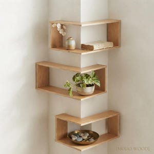 May include: Three light-coloured wooden corner shelves mounted in a vertical arrangement. The top shelf holds a small vase and a book, the middle shelf displays a potted green plant, and the bottom shelf contains a brass bowl filled with smooth stones.