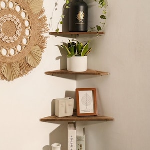 Op de afbeelding: Drie driehoekige houten hoekplanken gemonteerd aan een muur. De bovenste plank bevat een zwarte vaas met een gouden bladontwerp en klimop. De middelste plank toont een plant in pot. De onderste plank bevat een gezichtsvormige plantenbak.