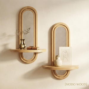 Peut inclure: Deux miroirs muraux arqués avec des cadres en bois clair, chacun doté d'une étagère semi-circulaire intégrée. Le miroir de gauche porte deux petits vases et un livre, tandis que celui de droite présente un dessin au trait encadré et un vase. MODO WOOD.