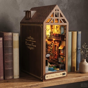 Puede incluir: Un inserto de estantería de madera con forma de casa, que presenta una escena callejera en miniatura iluminada de Colmar Town. El exterior marrón oscuro tiene la inscripción Welcome to fairy tale town Colmar Town y Colmar Town.