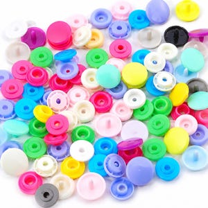 May include: A collection of assorted plastic snap fasteners in a variety of vibrant colours including pink, blue, green, yellow, white, purple, and black. These circular sewing components feature a raised centre post and a recessed underside.