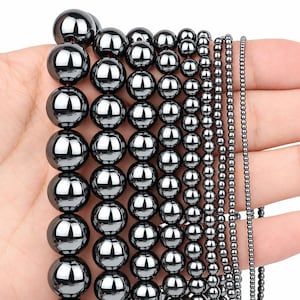 May include: A collection of smooth, spherical hematite beads arranged in multiple vertical strands of varying sizes, ranging from large, polished dark grey spheres on the left to tiny, uniform beads on the right, all featuring a high-gloss finish.
