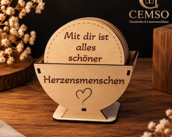 Può includere: Un set di sottobicchieri rotondi in legno chiaro in un supporto in legno abbinato. Il sottobicchiere superiore presenta il testo in tedesco 'Mit dir ist alles schöner', mentre il supporto è inciso con 'Herzensmenschen' e un piccolo cuore.
