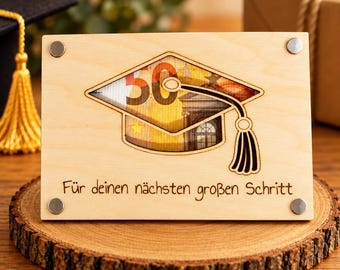 Può includere: Una targa rettangolare in legno chiaro con un intaglio a forma di tocco di laurea. Una banconota da cinquanta euro è visibile attraverso l'intaglio. Il testo "Für deinen nächsten großen Schritt" è inciso sotto il tocco.
