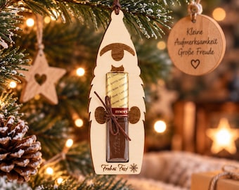 Può includere: Una decorazione in legno chiaro a forma di gnomo che sostiene una barretta di cioccolato Merci avvolta in carta dorata e bianca. L'ornamento è dotato di un cordino marrone per appenderlo e reca la scritta Frohes Fest con un fiocco di neve.