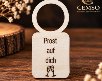 Può includere: Un'etichetta per bottiglie in legno chiaro con un'apertura circolare superiore. La sezione rettangolare inferiore presenta il testo inciso 'Prost auf dich' sopra una piccola grafica di due flûte da champagne che brindano.