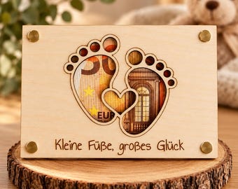 Può includere: Una targa rettangolare in legno chiaro con un intaglio a forma di due impronte di piedi di neonato e un cuore. L'intaglio rivela una banconota da cinquanta euro. Il testo "Kleine Füße, großes Glück" è inciso nella parte inferiore.