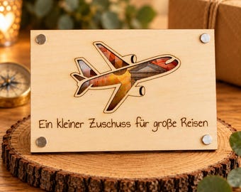 Può includere: Un cartello rettangolare in legno chiaro con un ritaglio a forma di aereo. Il ritaglio rivela un'immagine colorata di un edificio. Il testo "Ein kleiner Zuschuss für große Reisen" è inciso sotto la sagoma dell'aereo.