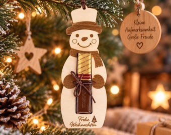 Può includere: Un ornamento in legno chiaro a forma di pupazzo di neve con cappello e sciarpa marrone scuro, che tiene una barretta di cioccolato rettangolare avvolta in carta dorata e marrone. La base reca l'incisione del testo Frohe Weihnachten e un piccolo albero.
