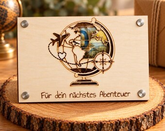 Può includere: Un salvadanaio rettangolare in legno chiaro con un design a globo tagliato al laser, un piccolo aereo, un cuore e una bussola. Il testo "Für dein nächstes Abenteuer" è inciso sul fondo. Quattro bulloni metallici argentati fissano il pannello frontale.