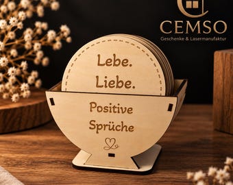 Può includere: Un set di sottobicchieri rotondi in legno chiaro riposti in un supporto in legno abbinato. I sottobicchieri presentano il testo inciso Lebe. Liebe. e il supporto mostra il testo Positive Sprüche con una piccola icona a forma di cuore.