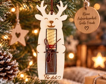Può includere: Un ornamento a forma di renna in legno chiaro con un ritaglio centrale che contiene una barretta di cioccolato legata con uno spago marrone. L'ornamento è inciso con il testo tedesco "Für dich" e un cuore, appeso a un cordino marrone.