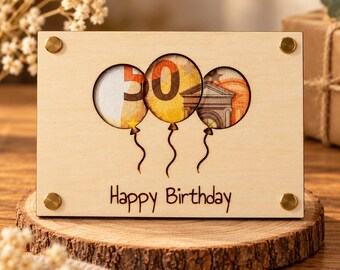 Può includere: Una targa rettangolare in legno chiaro con tre ritagli circolari a forma di palloncino. All'interno dei ritagli è visibile una banconota da 50 euro. Il testo "Happy Birthday" è inciso sotto i palloncini. Quattro borchie in metallo color ottone sono fissate agli angoli della tavoletta.