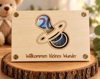 Può includere: Una targa rettangolare in legno chiaro con un ritaglio a forma di ciuccio, con una banconota da venti euro visibile dietro l'apertura. Il testo "Willkommen kleines Wunder" è inciso nella parte inferiore, con quattro fissaggi metallici color ottone agli angoli.