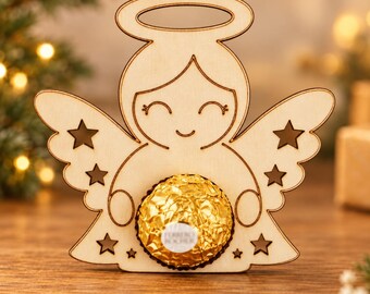 Può includere: Una sagoma di angelo in legno chiaro con aureola, tratti del viso incisi e ritagli a forma di stella sulle ali. Un cioccolatino Ferrero Rocher rotondo, avvolto in carta dorata, è posizionato al centro del corpo dell'angelo.