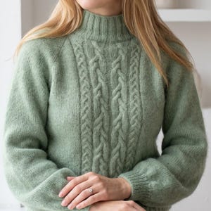 May include: A sage green long-sleeve jumper featuring a high mock neck collar and a central vertical cable knit pattern. The garment is crafted from a soft, textured knit fabric with ribbed cuffs and a ribbed neckline, providing a fitted silhouette.