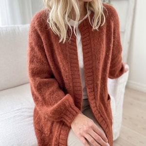 May include: A rust-colored open-front cardigan featuring a textured knit pattern, long sleeves with ribbed cuffs, and a wide ribbed placket along the front opening. The garment is worn over a plain white shirt and light-colored trousers.