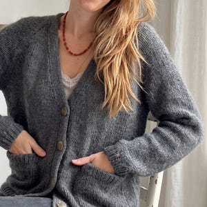 May include: A charcoal grey knit cardigan featuring a V-neckline, long sleeves, and two front patch pockets. The garment is fastened with round buttons down the centre front and is worn over a white camisole with lace trim and a beaded necklace.