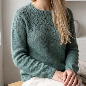 May include: A sage green long-sleeve jumper featuring a round neckline and a detailed knit pattern of vertical leaf-like motifs across the upper chest and shoulders. The garment has ribbed cuffs and a ribbed hem, with a solid knit texture below.