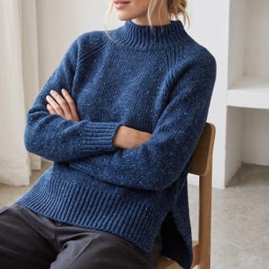 May include: A long-sleeved, dark blue speckled sweater featuring a mock neck collar, ribbed cuffs, and a ribbed hemline. The knit garment has a high-low hem design with side slits, showcasing a textured, flecked pattern throughout the fabric.