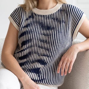 May include: A sleeveless knit sweater vest featuring a vertical striped pattern in navy blue and cream. The garment has a round ribbed neckline and wide armholes, constructed with a textured ribbed knit fabric throughout the entire piece.