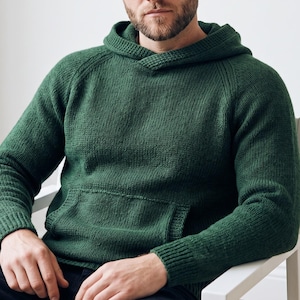May include: A forest green knit hoodie featuring a textured hood, long sleeves with ribbed cuffs, and a large front kangaroo pocket. The garment is constructed with a visible knit stitch pattern and raglan sleeves, providing a consistent solid colour.