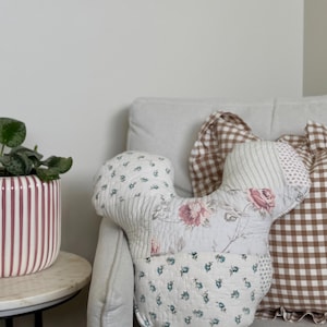May include: A patchwork throw pillow shaped like a mouse head, featuring quilted fabric with floral patterns in blue, pink, and white. The pillow is constructed from various fabric segments, including small blue floral prints, striped sections, and larger pink rose motifs.
