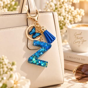 May include: A gold-toned keychain featuring a blue butterfly charm, a blue tassel, and a letter Z pendant filled with blue and teal glitter. The keychain is attached to a beige handbag. The letter Z is crafted from resin with various shades of blue.