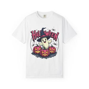 Op de afbeelding: Een wit t-shirt met een afbeelding van een lachende geest met een heksenhoed, omringd door drie pompoenen met gloeiende gele ogen en monden. Het woord HALLOWEEN staat in het roze boven de geest gedrukt, samen met kleine zwarte spinnen.