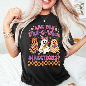 May include: A black t-shirt featuring a graphic with the text ARE YOU FALL-O-WEEN DIRECTIONS? The design includes three ghosts: one with a leopard print, one with a pink polka dot bow holding a pencil, and one with an orange checkerboard pattern.